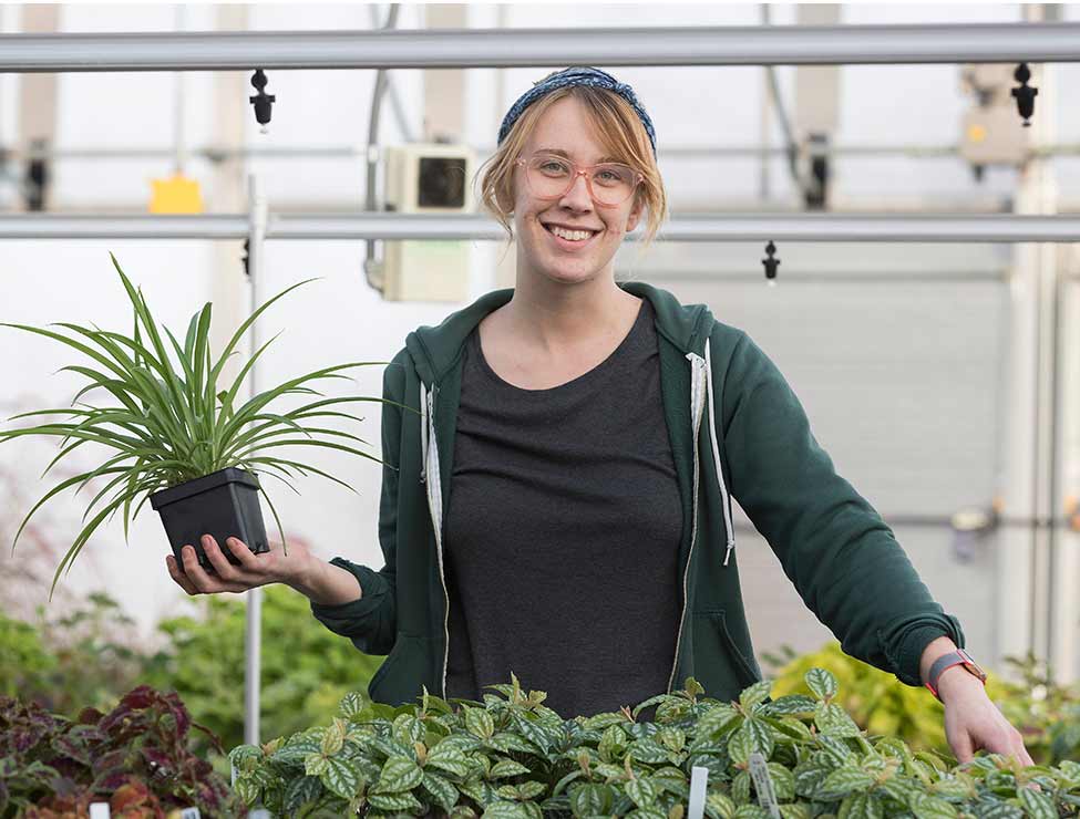 Student in green sweatshirt smiling with plant in greenhouse